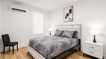A bedroom with a bed, bedside tables, a chair, an air conditioner, and a window with blinds.