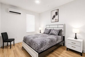 Quarto com cama, mesas de cabeceira, uma cadeira, ar-condicionado e janela com persianas.