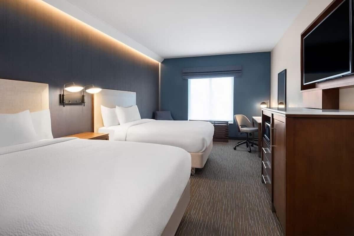 Ein Hotelzimmer mit zwei Betten, einem Schreibtisch mit Stuhl, einem Fernseher und einem großen Fenster.