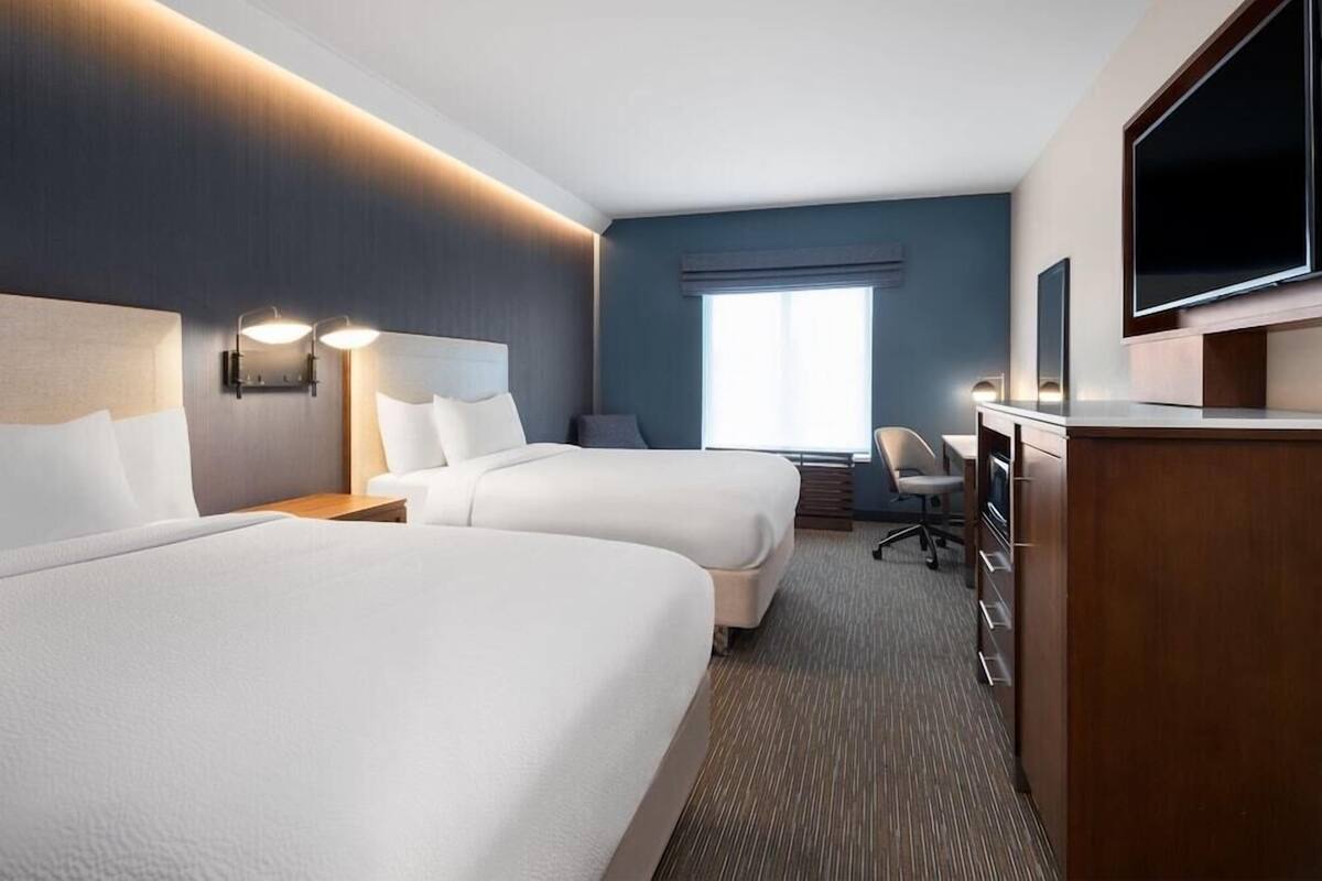 Ein Hotelzimmer mit zwei Betten, einem Schreibtisch mit Stuhl, einem Fernseher und einem großen Fenster.