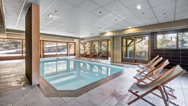 Une piscine intérieure moderne avec des parois en verre transparent, des chaises longues en bois et de grandes fenêtres offrant une vue sur l’extérieur.