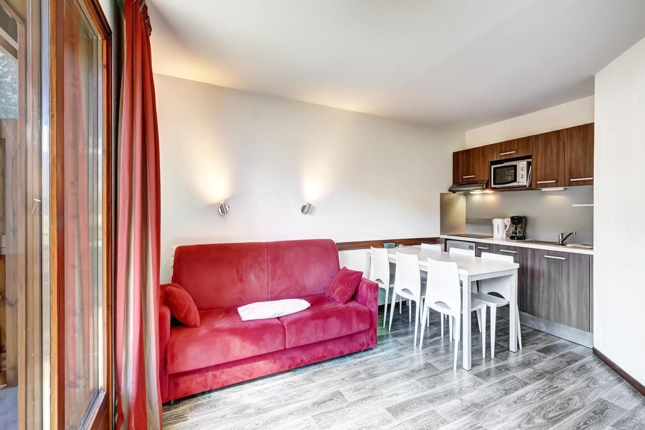 A modern living room with a red sofa, white dining table, and chairs. A kitchen area with wooden cabinets and a microwave is visible.