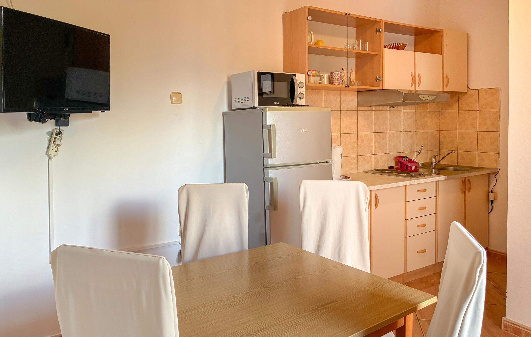 Cozinha compacta com micro-ondas, frigorífico e uma pia pequena. Há uma mesa de jantar com cadeiras e uma televisão fixada na parede.