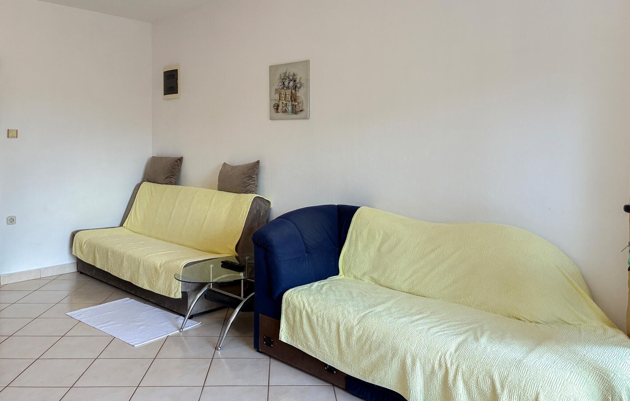 Une pièce avec deux canapés, un jaune et un bleu, une table basse en verre et un petit tableau accroché au mur.