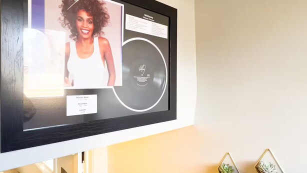 Un retrato enmarco de Whitney Houston en una pared, con fecha de lanzamiento y detalles de grabación.