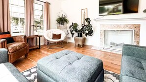A living room with a fireplace, a television mounted on the wall, and a large window with curtains. - Amazing 3-Bed Cottage with Spa, Home Office & Gym (Idaho Falls)