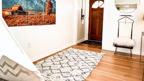 A hallway with a wooden floor, a white rug, a wooden door, a mirror, and a wall-mounted shelf. - Amazing 3-Bed Cottage with Spa, Home Office & Gym (Idaho Falls)