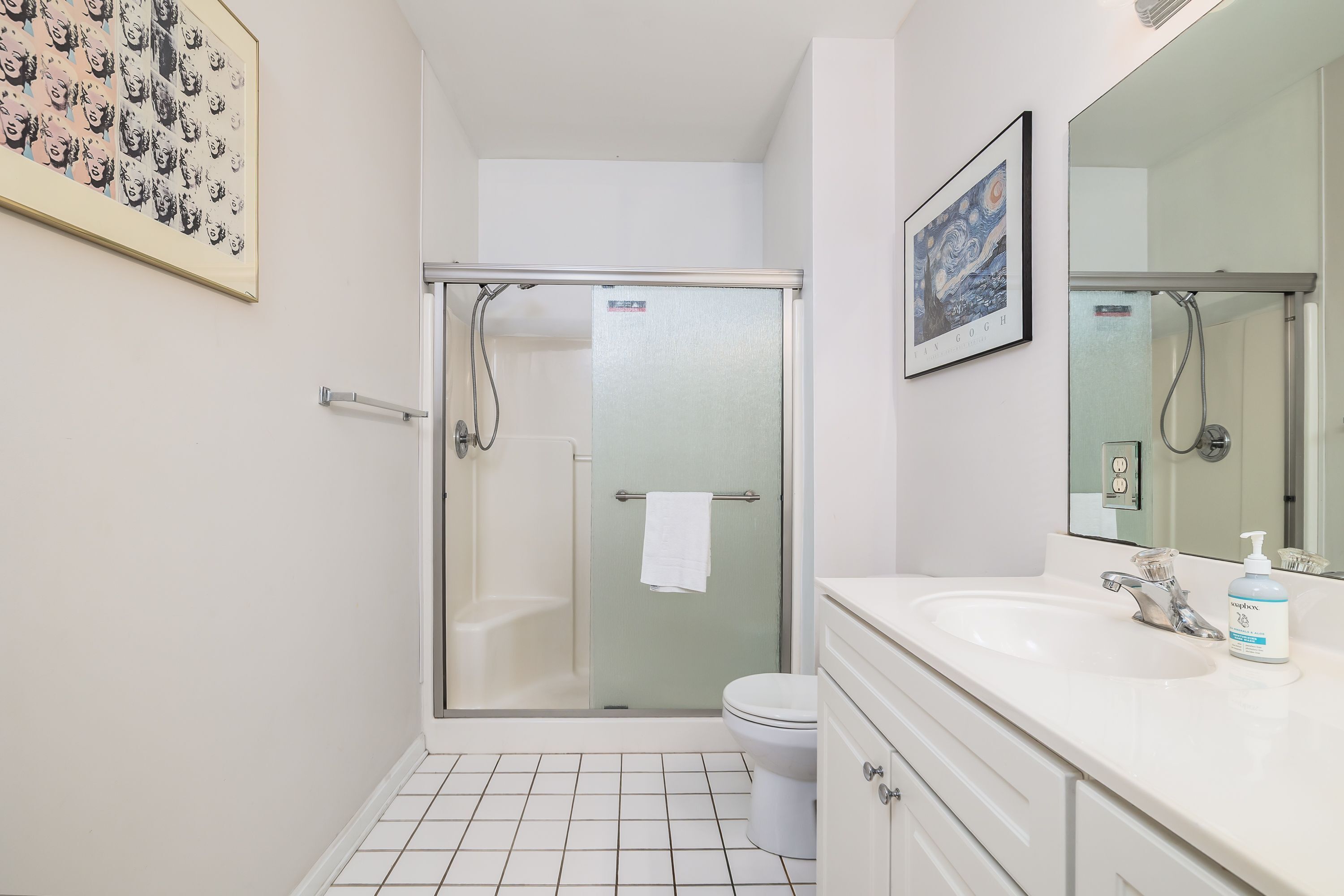 A bathroom with a glass-enclosed shower, a toilet, a sink with a mirror, and a framed artwork on the wall.
