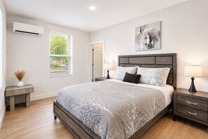 A bedroom with a large bed, bedside tables, an air conditioning unit, a window with blinds, and a painting on the wall. - Elegant 2 Bed 2 Bath • Downtown • Free Parking (Hot Springs)