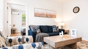 Un salon moderne avec un canapé, un fauteuil et une table basse. Un tableau est accroché au mur, et une horloge est visible.