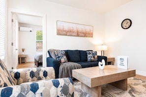 A modern living room with a sofa, armchair, and coffee table. A painting hangs on the wall, and a clock is visible. - Elegant 2 Bed 2 Bath • Downtown • Free Parking (Hot Springs)