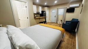 Een slaapkamer met een bed, nachtkastje en stoel. Er is een kitchenette met een magnetron en een koelkast.