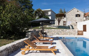 A poolside area with wooden lounge chairs, a stone wall, and a building with a balcony. - Amazing home in Donji Bitelic (Donji Bitelic)