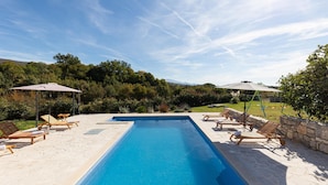 Una piscina rectangular con borde de piedra, rodeada de sillas de descanso y sombrillas.