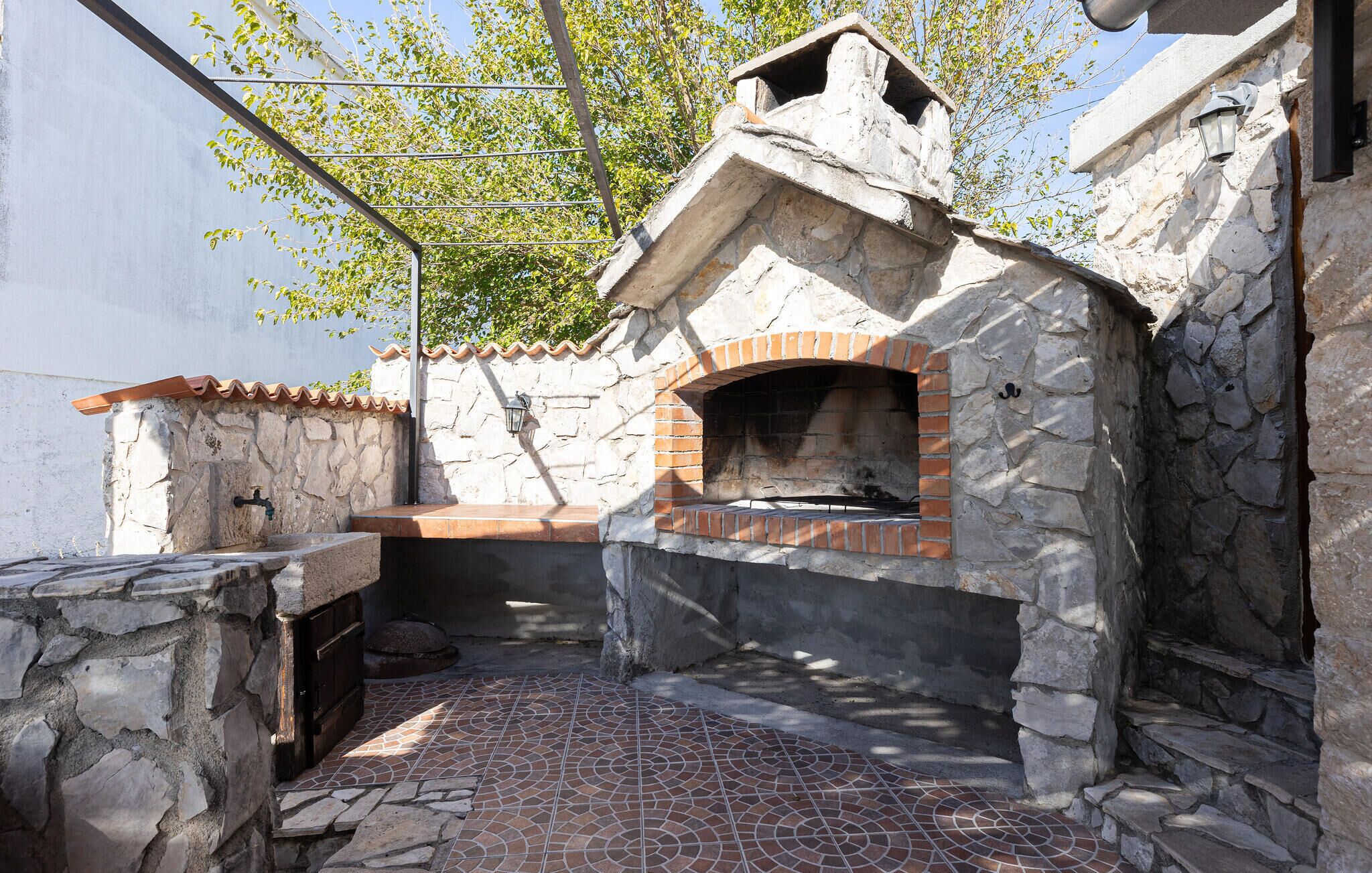 A stone-built outdoor kitchen with a brick oven, a tiled floor, and a metal roof structure.