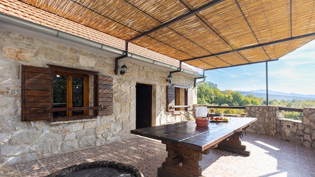 Un edificio de piedra rústico con una mesa y sillas de madera bajo un techo de paja, con vistas a un paisaje montañoso.