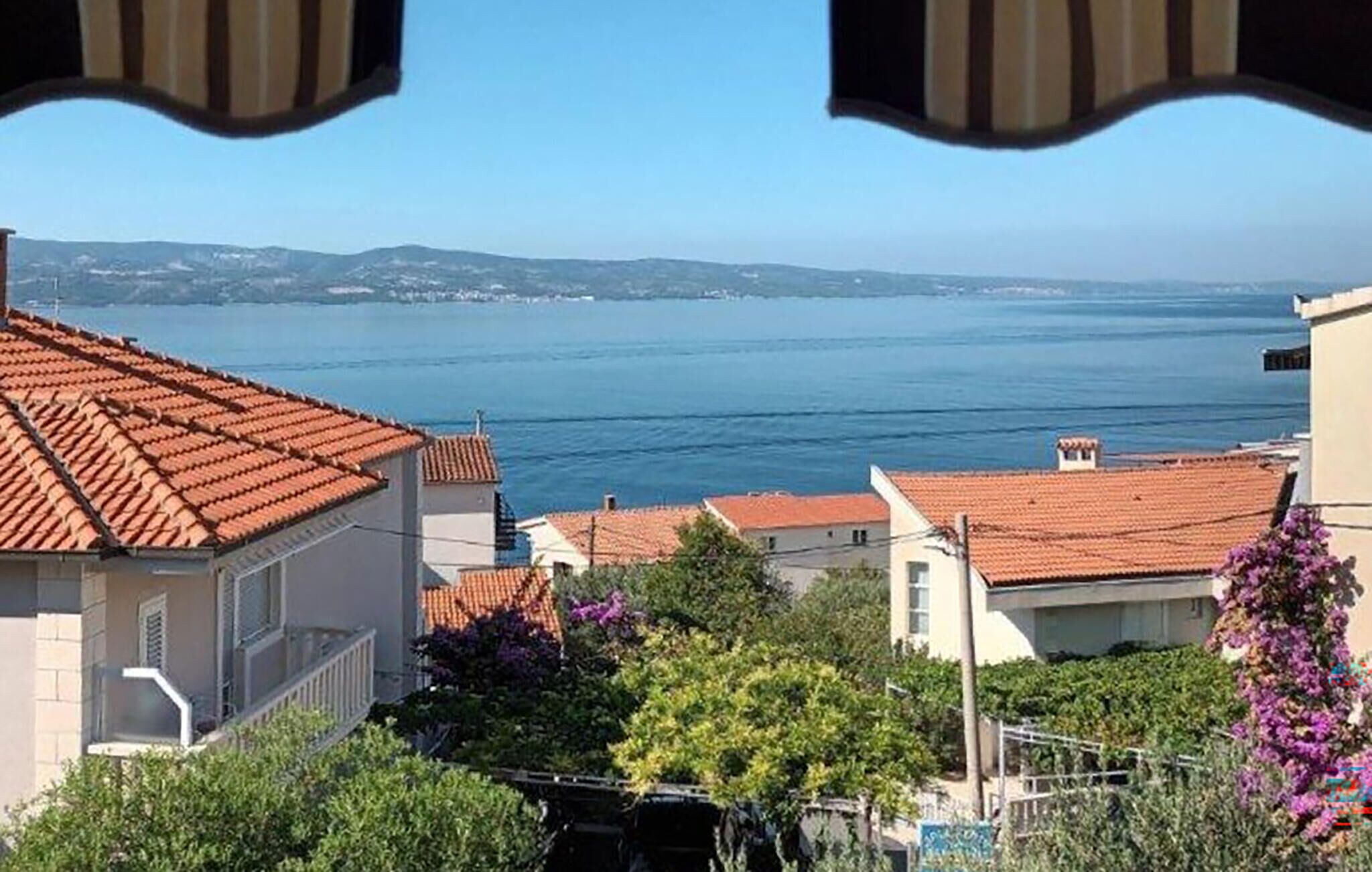 A coastal view with terracotta-roofed buildings, a clear sky, and a calm sea.