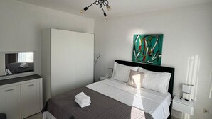 Et hotelværelse med seng, garderobe, skrivebord med stol, et billede på væggen og en moderne lampe.