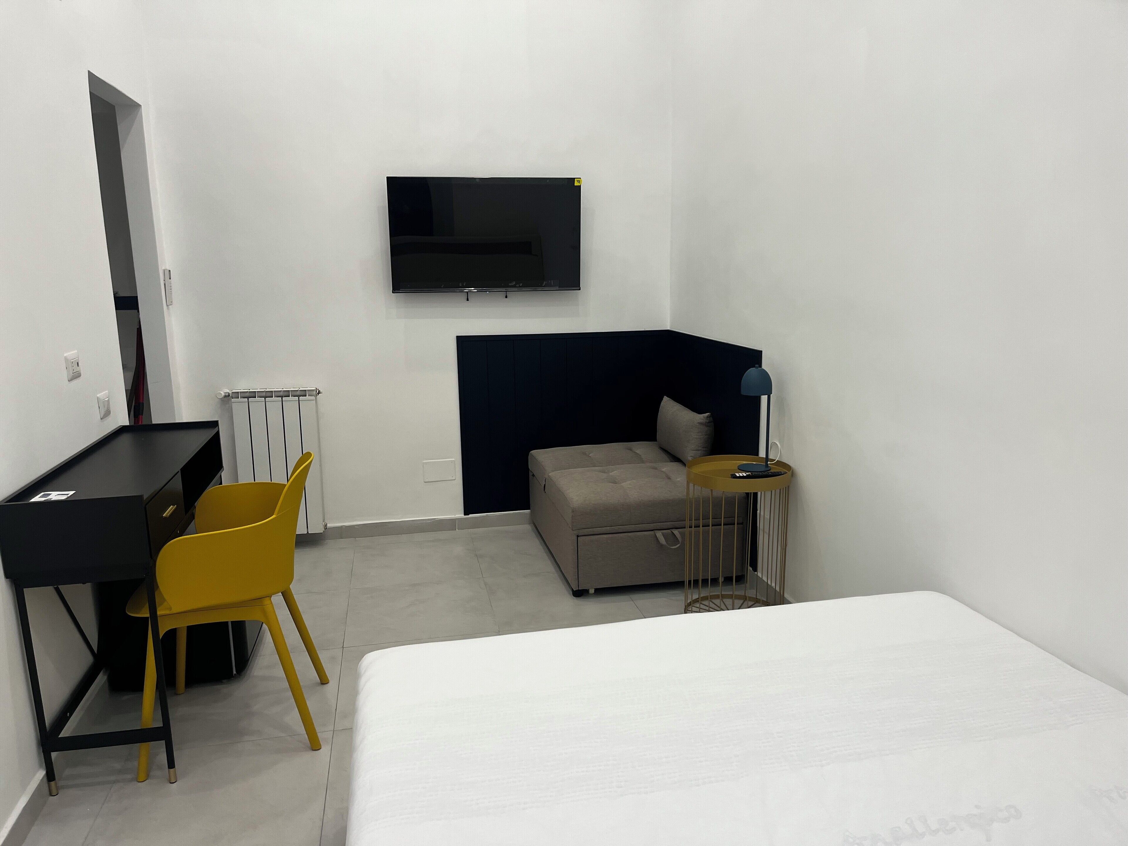 Habitación de hotel con cama, escritorio, silla, televisor montado en la pared y mesita de noche con lámpara.