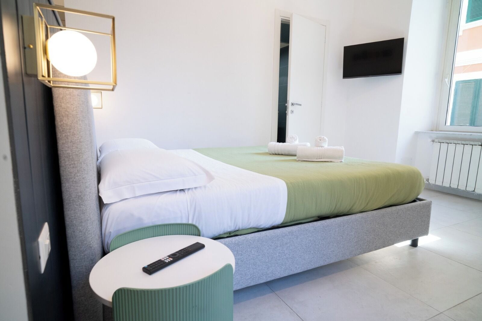 Une chambre d’hôtel moderne, équipée d’un lit, d’une petite table ronde, d’un téléviseur fixé au mur et d’une fenêtre avec des stores.