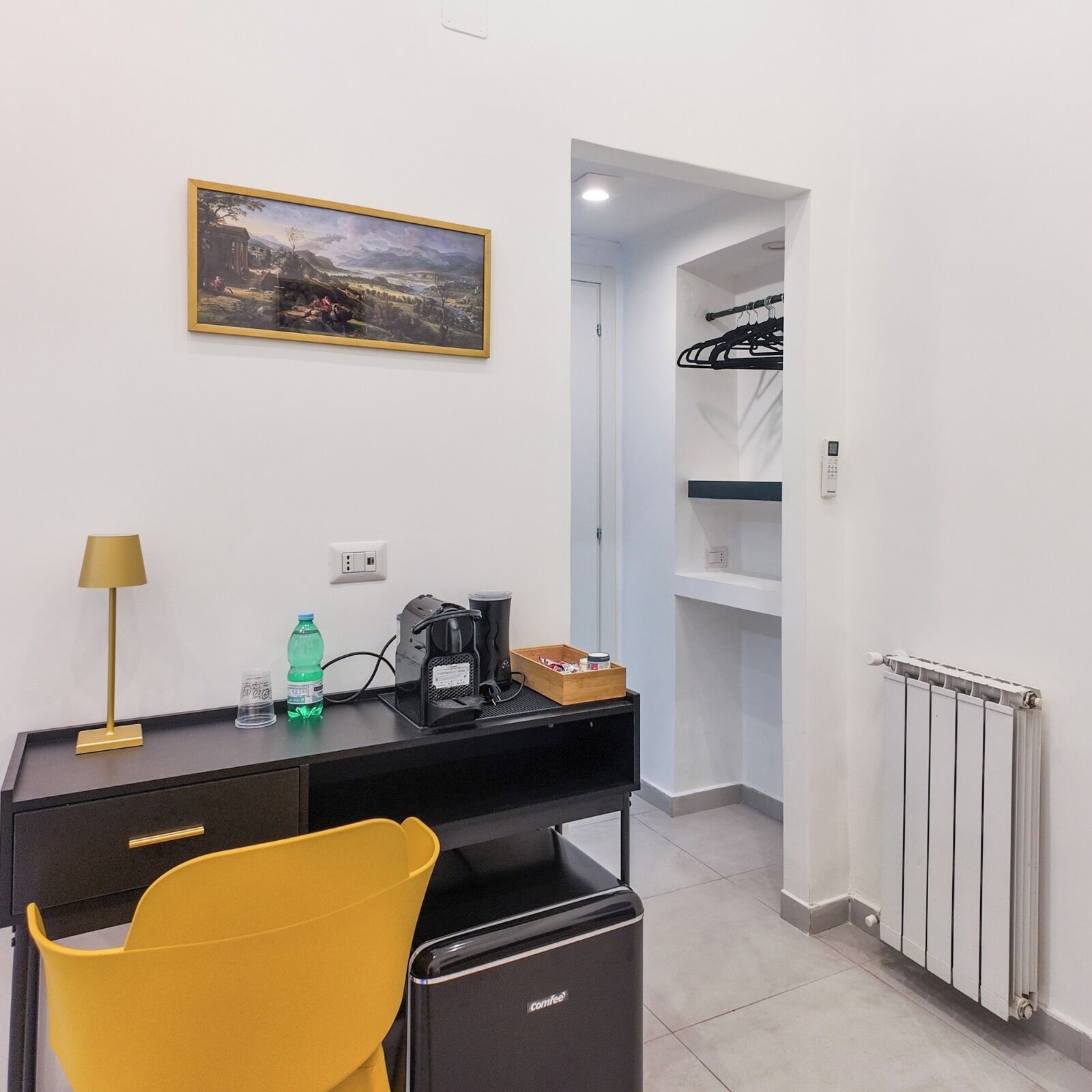 Een hotelkamer met een bureau, een gele stoel, een koffieapparaat, een fles water, een lijst met foto aan de muur en een inloopkast met hangende kleding.