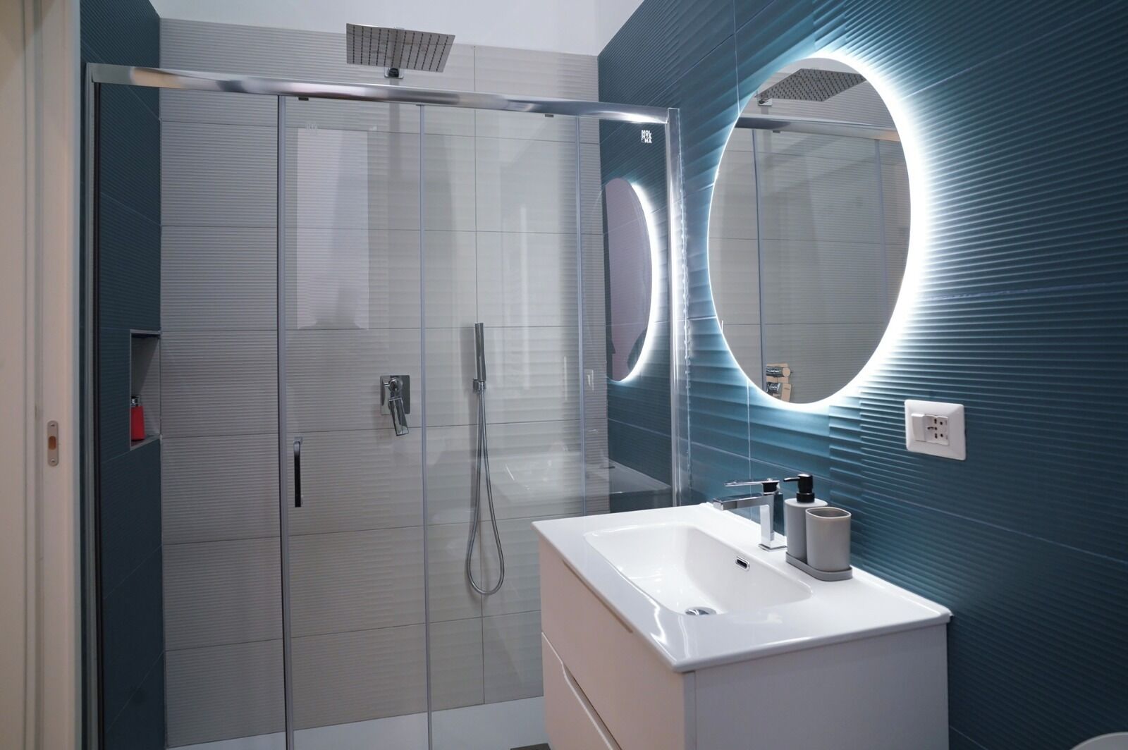 A modern bathroom with a glass-enclosed shower, a white vanity, and a large mirror with a circular light.