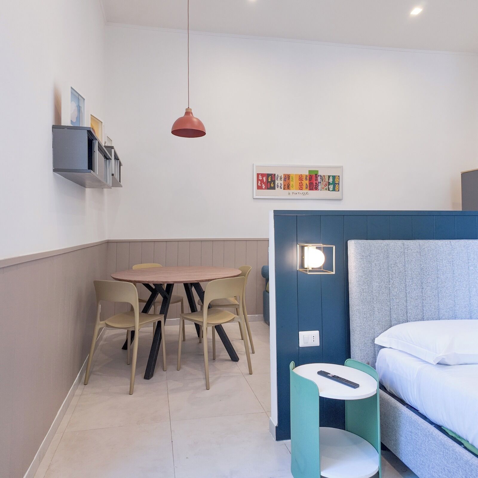 A compact room with a bed, a small table with chairs, a hanging red pendant light, a wall-mounted shelf with books, and a framed artwork.