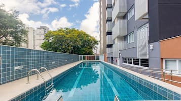 Piscina com grades de metal, parede revestida e uma árvore ao fundo.
