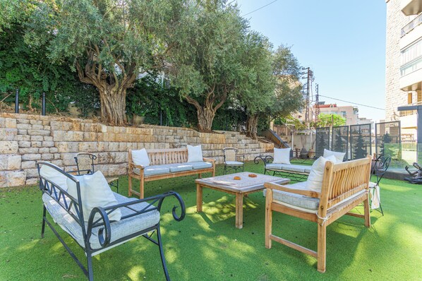 An outdoor seating area with a stone wall, wooden furniture, and trees. - GlassHaus Rooftop 2-BR & Pool in Ghazir (Ghazir)