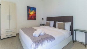 A neatly made bed with a white comforter and a folded towel, a brown headboard, a bedside table with a lamp, and a beige wardrobe. - GlassHaus Rooftop 2-BR & Pool in Ghazir (Ghazir)