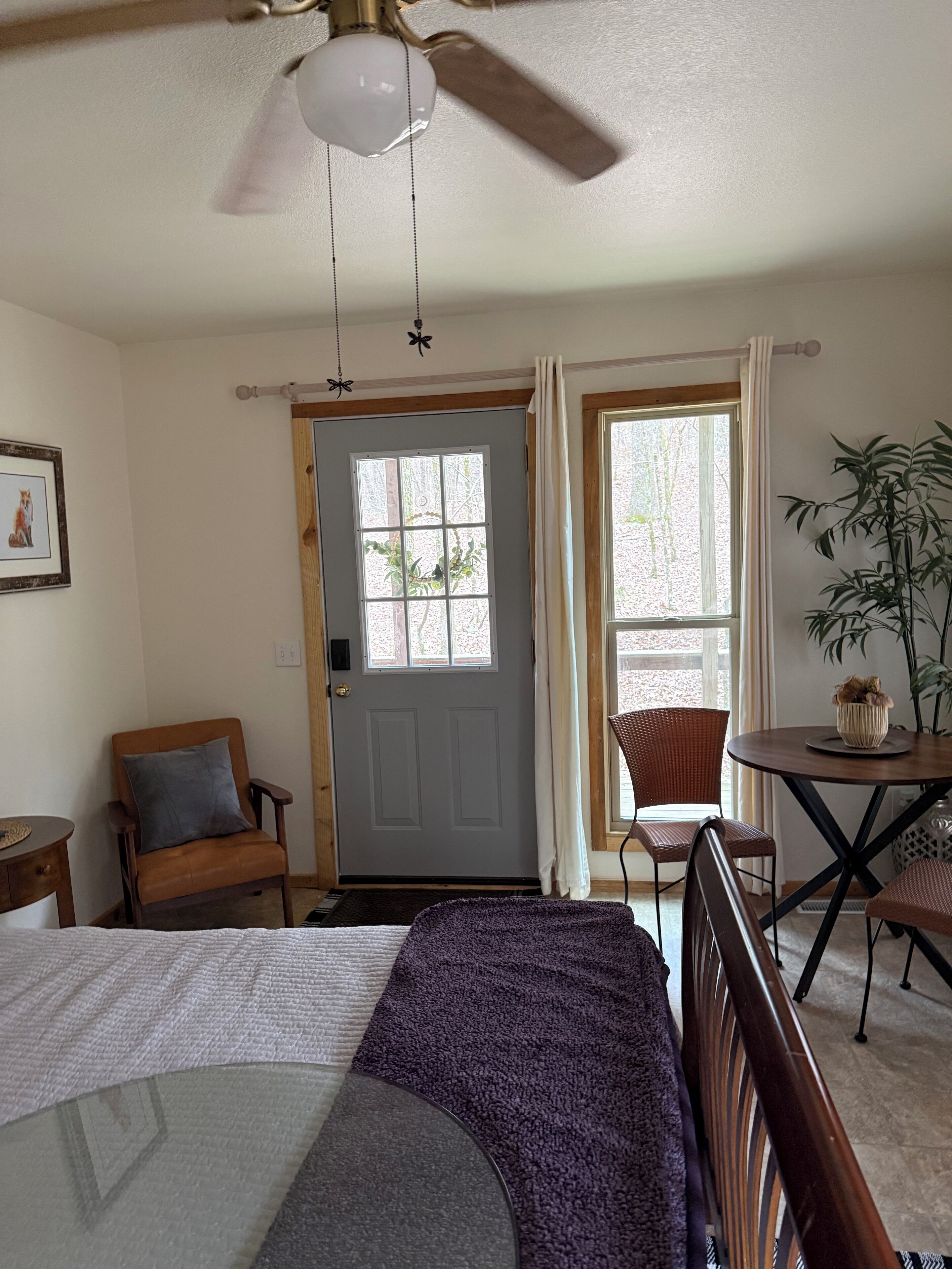 A room with a bed, a chair, a table, a ceiling fan, a door, and a window.