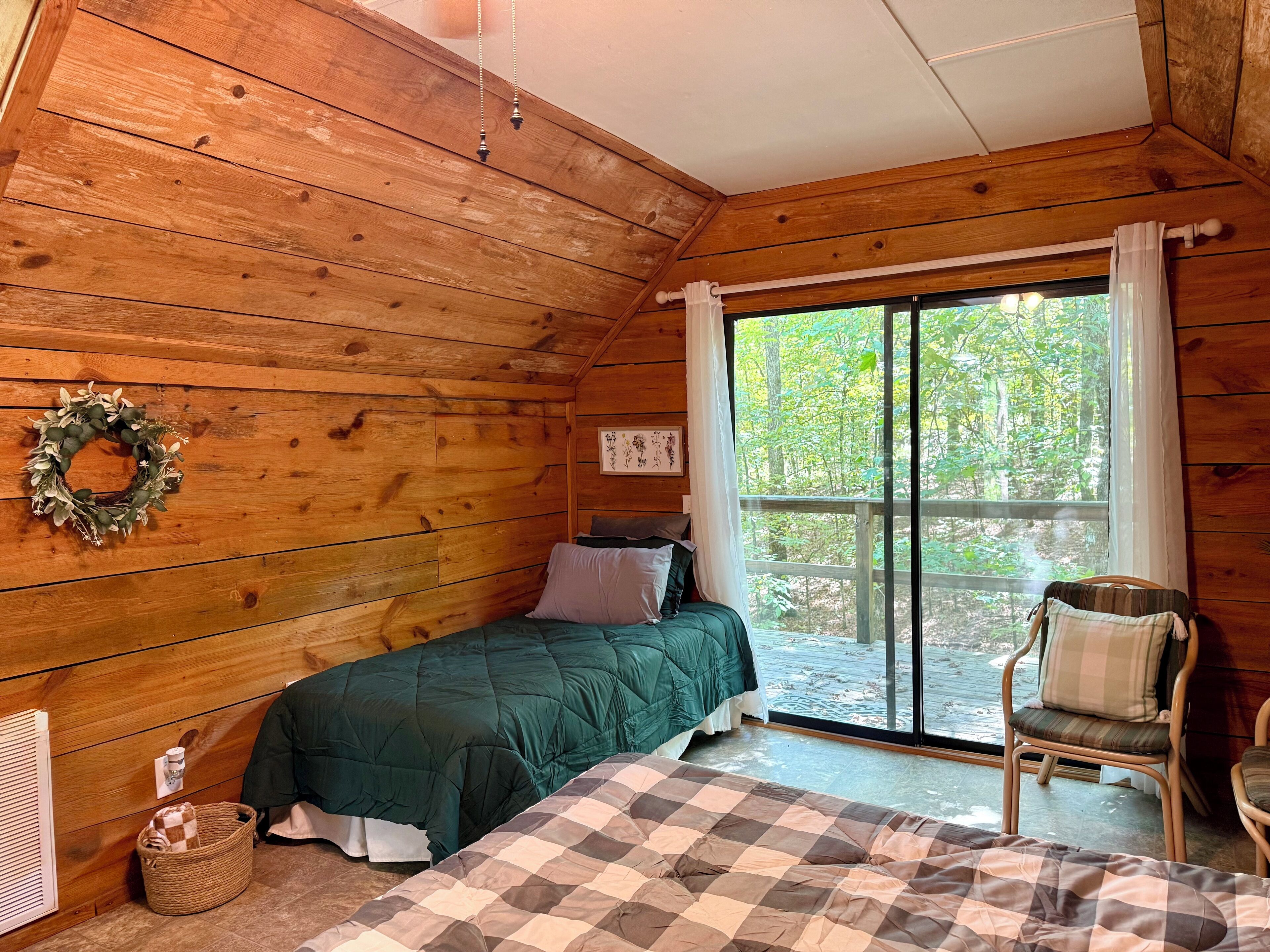 Une pièce chaleureuse dans un chalet, avec des murs en bois, un lit recouvert d’une couverture verte et d’une courtepointe à carreaux, un panier en osier et une chaise placée près d’une grande fenêtre offrant une vue sur les arbres.