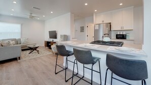 Una cucina moderna con mobili bianchi, un frigorifero in acciaio inossidabile e un piano cottura a gas. C'è un'area pranzo con uno sgabello da bar e un divano.