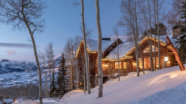 A log cabin with a warm-lit interior, surrounded by snow-covered trees and a mountainous backdrop. - The Wilson Vista: Epic Space & Solitude (Mountain Village)