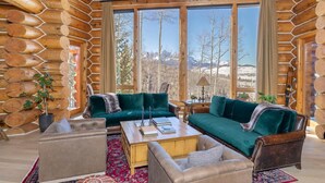 A log cabin interior with a green sofa set, leather armchairs, and a wooden coffee table. Large windows offer a view of a snowy landscape. - The Wilson Vista: Epic Space & Solitude (Mountain Village)