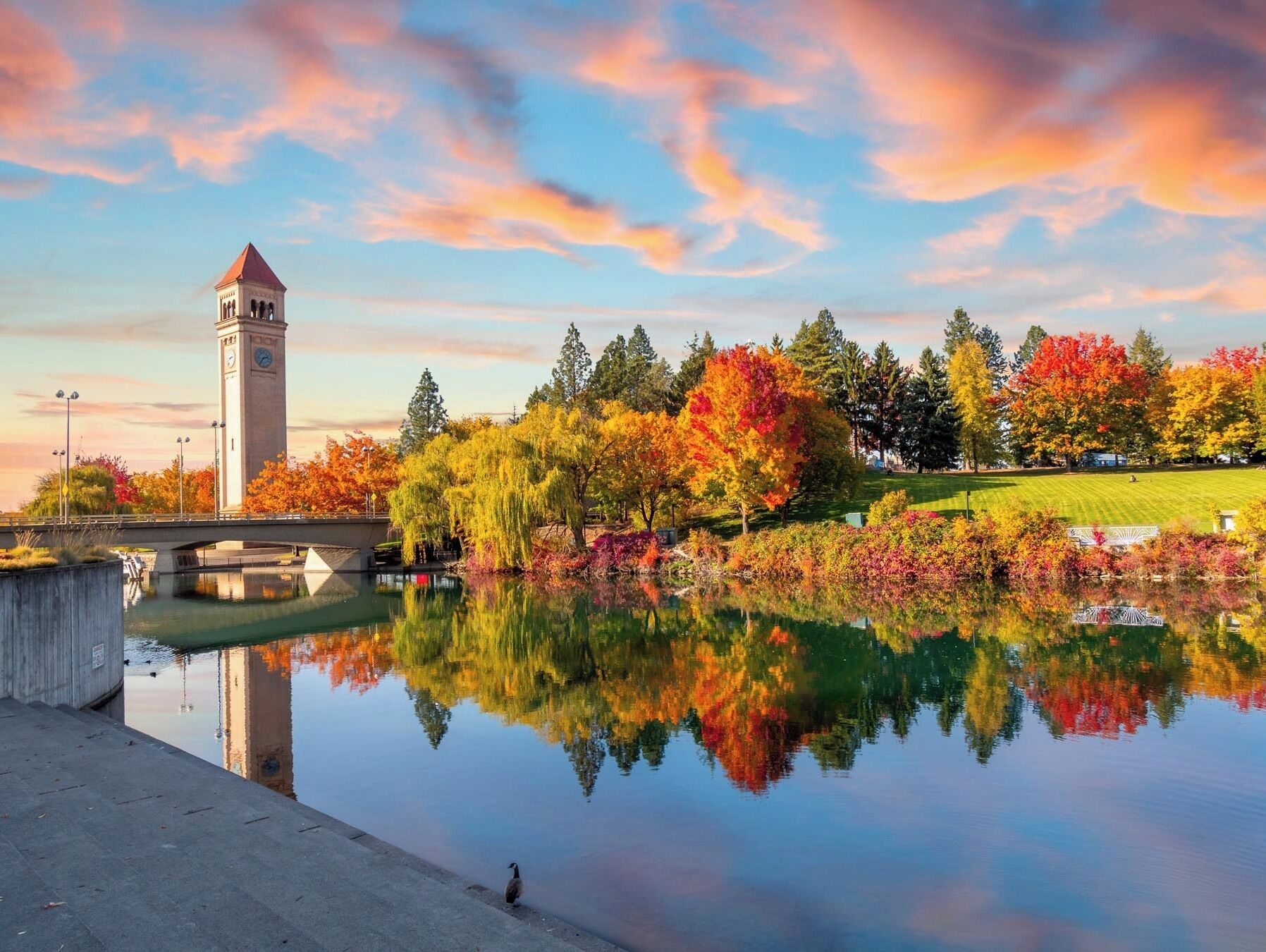 A park with a clock tower, a bridge, and a calm pond reflecting colorful trees.