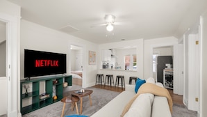 Una sala de estar moderna con un televisor de pantalla plana mostrando 'NETFLIX', un sofá, una mesa de centro y una cocina al fondo.