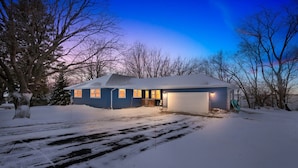 A snow-covered house with a garage, surrounded by trees, during twilight. - Lakefront Retreat w/ Dock + Games (Madison Lake)
