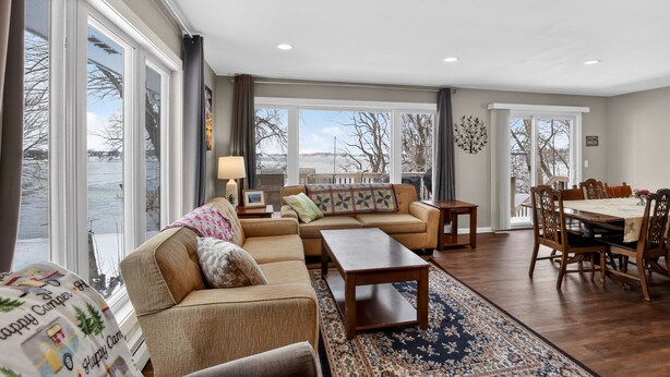 A spacious living room with a large window, a sofa, a coffee table, and a dining area with a table and chairs. - Lakefront Retreat w/ Dock + Games (Madison Lake)
