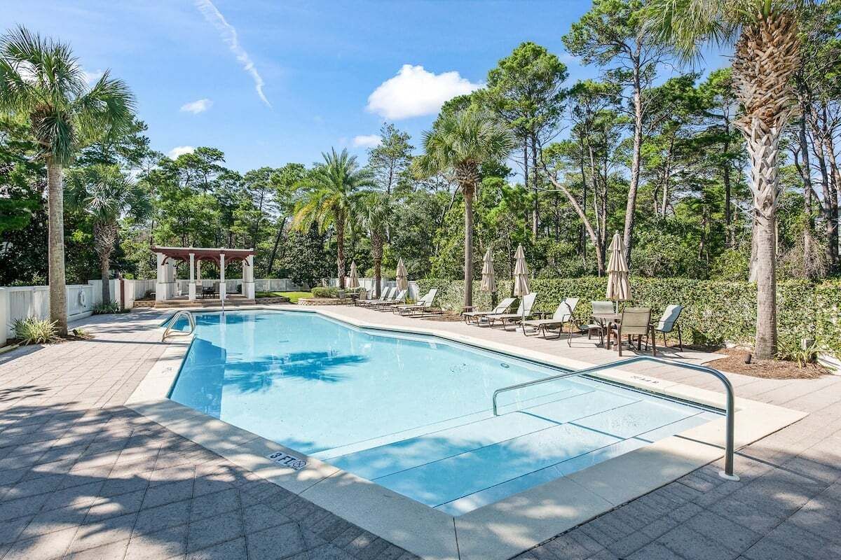 Una piscina rectangular con bordeado de baldosas, rodeada de sillones de descanso y sombrillas, con un pabellón y altos árboles al fondo.
