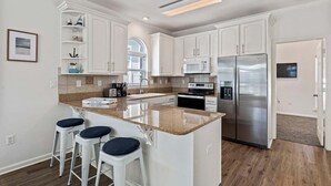 A modern kitchen with white cabinets, a stainless steel refrigerator, and a granite countertop. There are three white bar stools with blue cushions. - Private Pool, LSV/Golf Cart, Bikes, & Pet Friendly! 1/2 MILE TO BLUE MOUNTAIN BEACH! (Santa Rosa Beach)