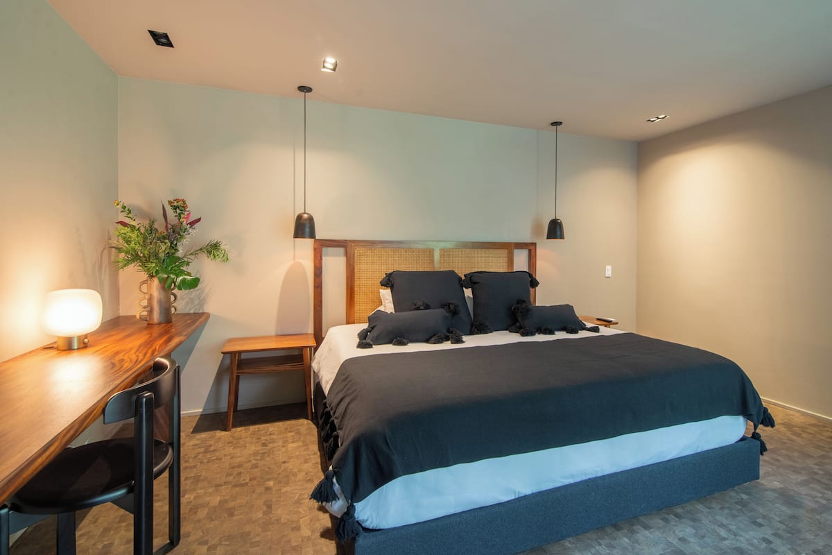 A modern hotel room with a large bed, a desk with a chair, a nightstand, and a vase with flowers.