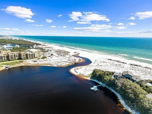 An aerial view of a coastal area with a large body of water, a sandy beach, and several buildings including a hotel with multiple floors and a dock extending into the water. - Steps to Beach! Private Pool, 6-Seater LSV/golf cart, & 4 Bikes Included! Sleeps 18! (Santa Rosa Beach)