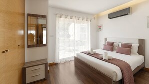 A modern bedroom with a wooden wardrobe, a large bed with a headboard, a bedside table, and a window with sheer curtains.