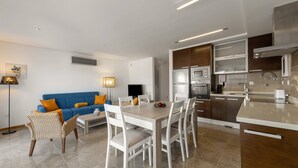 A modern kitchen with white cabinetry, a dining area with a wooden table and white chairs, and a blue sofa with orange cushions.