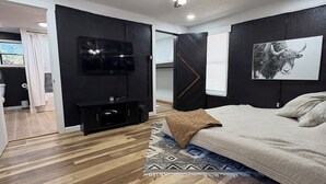 Une chambre moderne avec un plancher en bois, un lit, un ventilateur de plafond, une télévision et une œuvre d’art en noir et blanc.