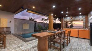 Un espace repas avec une piscine, des meubles en bois et une vue sur le coucher du soleil.