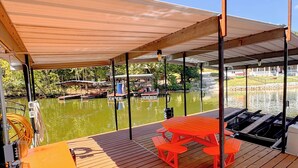 A covered deck with a picnic table and orange storage box, overlooking a body of water with boats and a dock in the background. - Lakefront Retreat - Mile Marker 42 - Dock Bar + Sleeps 15! (Roach)