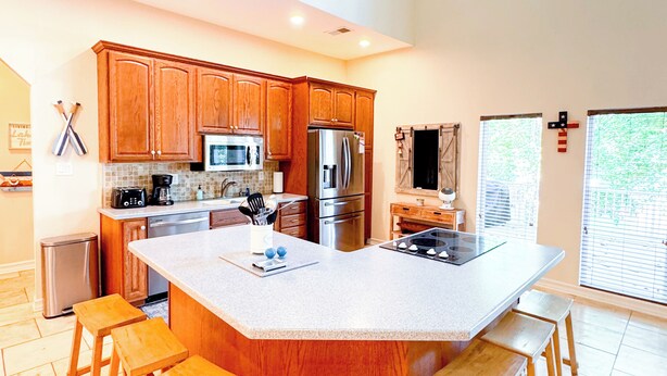 A spacious kitchen with wooden cabinets, a large island with stools, stainless steel appliances, and a view of the outdoors through the windows. - Lakefront Retreat - Mile Marker 42 - Dock Bar + Sleeps 15! (Roach)