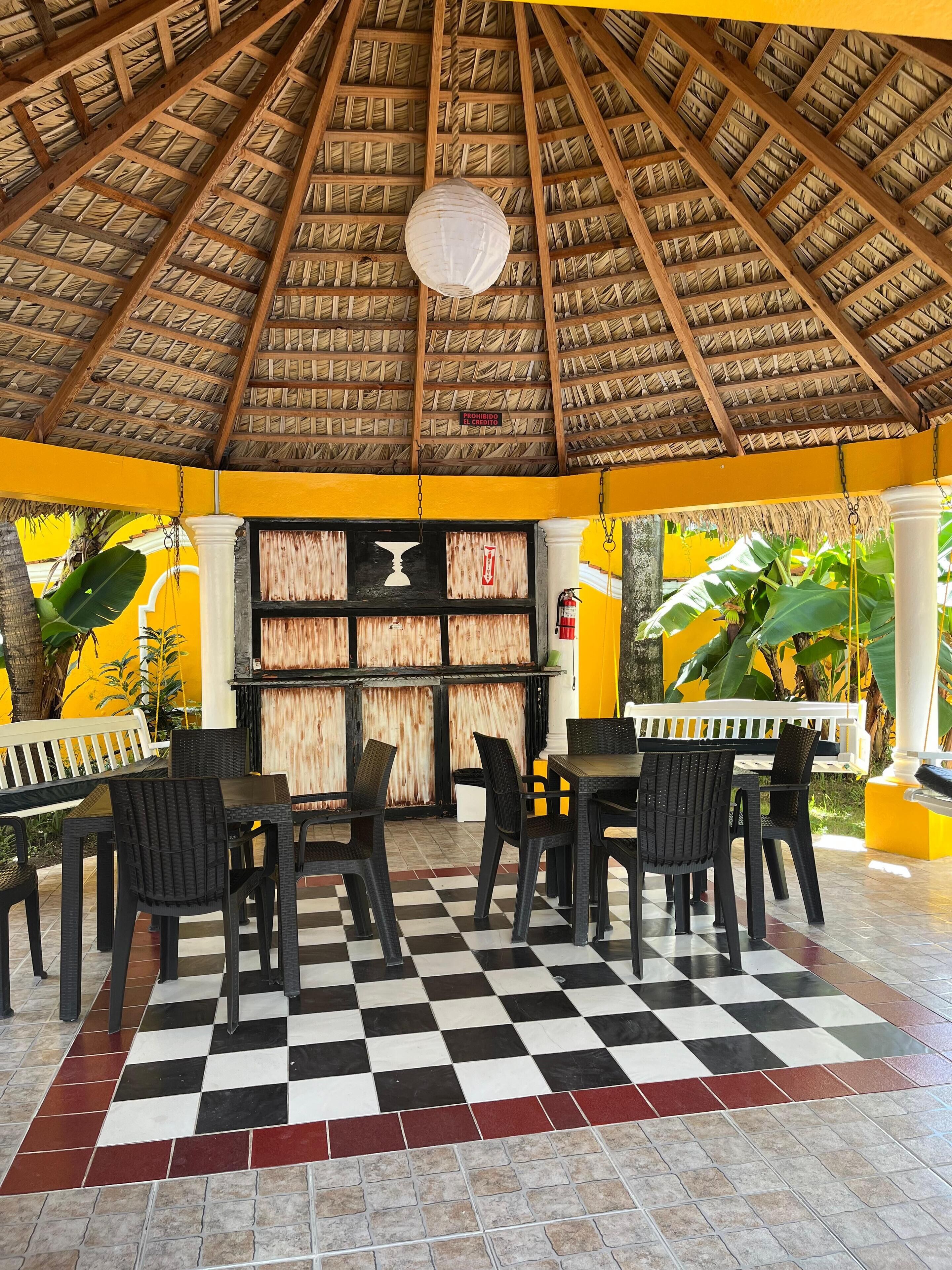 A covered outdoor seating area with a checkered floor, wooden beams, and a thatched roof.